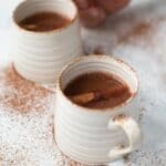 a closeup of this homemade Christmas Hot Chocolate in two white coffee mugs garnished with cacao powder and cinnamon sticks.