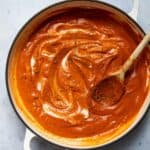Tomato soup in a large pot with a wooden spoon. Pot is displayed on a white marble background.