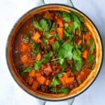 Sweet Potato and Black Bean Stew served in a large pot topped with coriander. The pot is displayed on a white marble background.