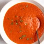 red pepper and tomato soup served in a white bowl with a spoon inside.