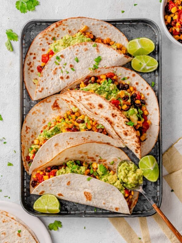 Sweetcorn and black bean tacos with avocado smash on a tray with lime wedges