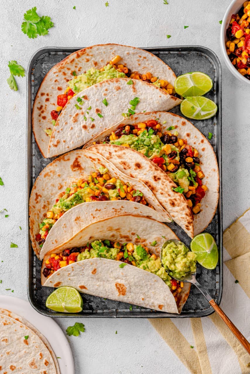 Sweetcorn and black bean tacos with avocado smash on a tray with lime wedges
